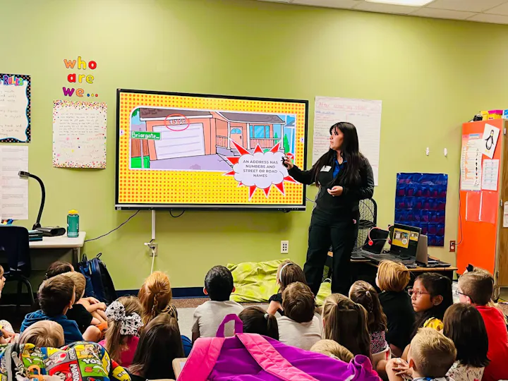 A teacher presents to a group of children, explaining an address using a cartoon on a screen in a classroom setting.