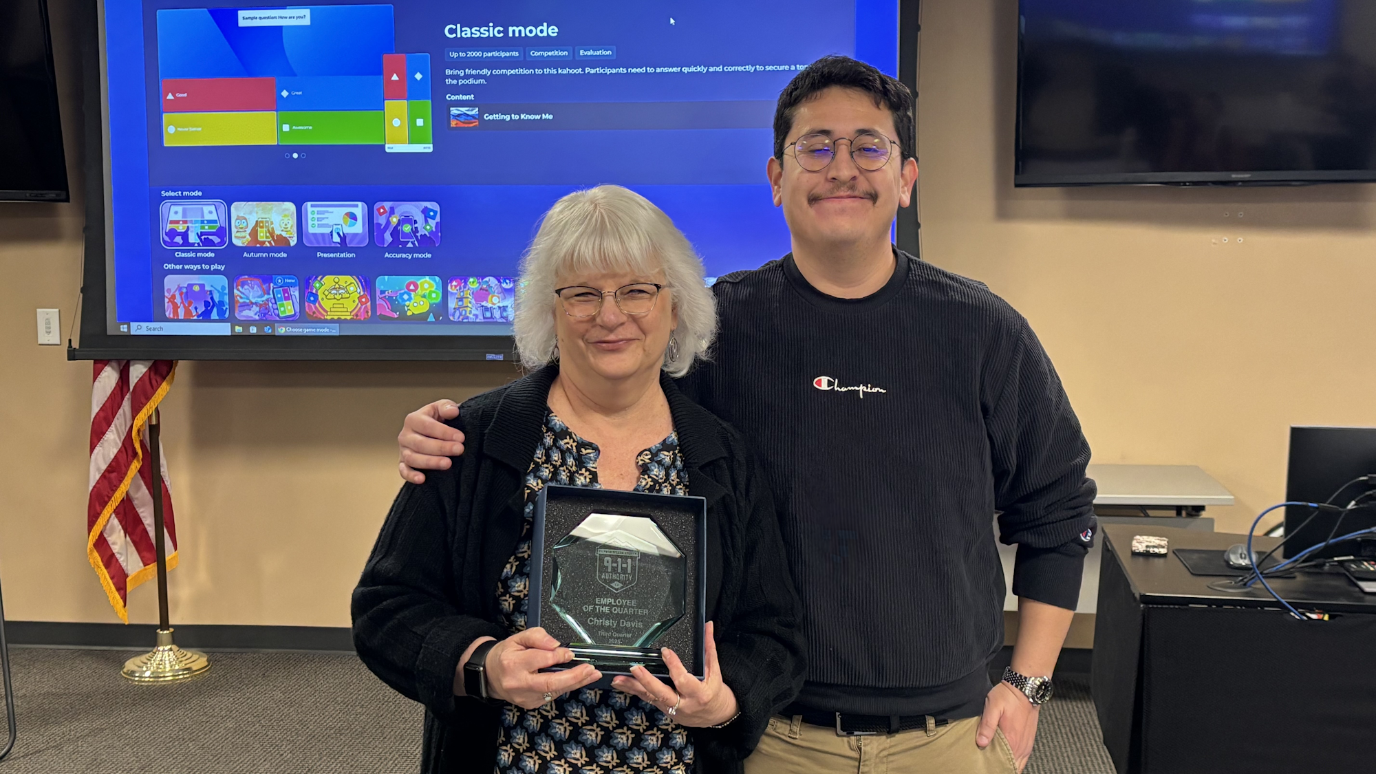 A woman and a man pose together, celebrating her "Employee of the Quarter" award in a professional setting with a screen behind them.