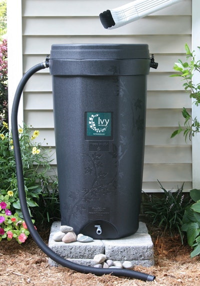 The image shows a dark rain barrel with a hose, positioned among flowers and plants beside a house.