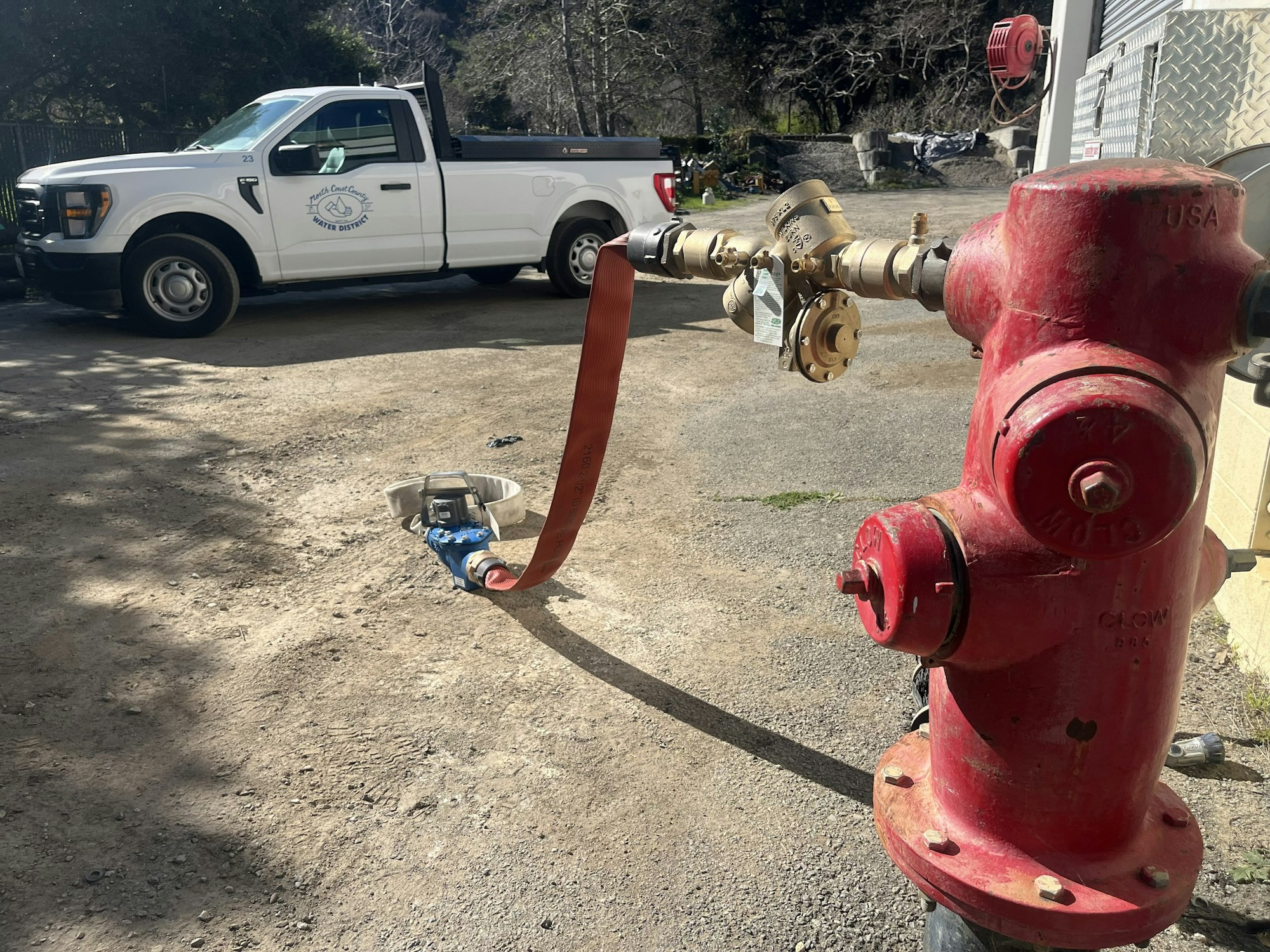 A red fire hydrant with a hose attached, alongside a white truck labeled "Water District," parked on a gravel surface.