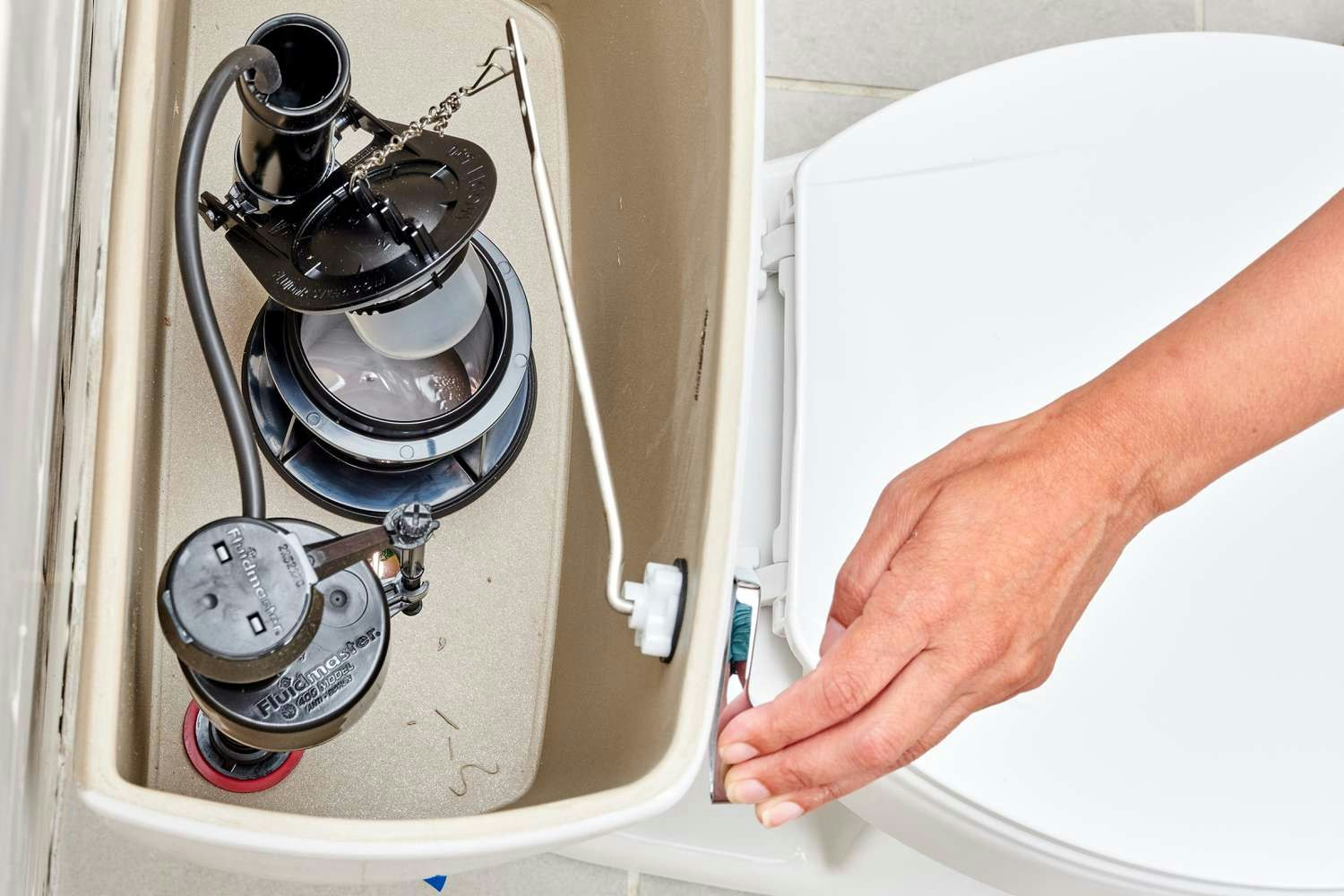 Inside a toilet tank, showing the flapper, flush mechanism, and a hand adjusting the water supply.