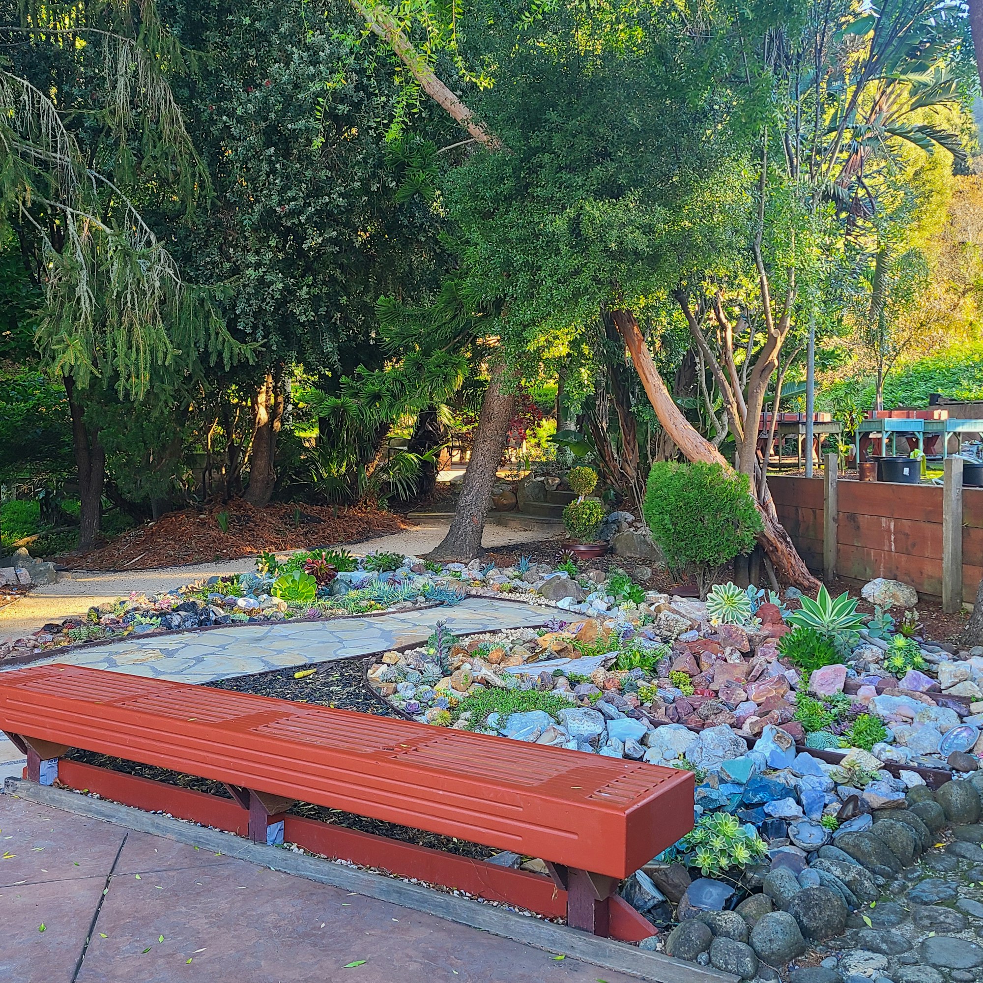 A lush garden scene featuring greenery, colorful rocks, a stone path, and a red bench, creating a serene outdoor space.