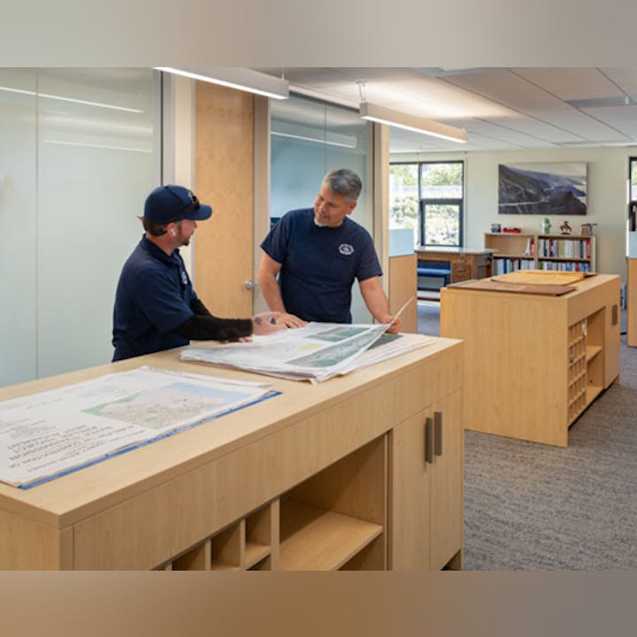 Two men examine large printed maps in an office setting with wooden furniture and large windows.