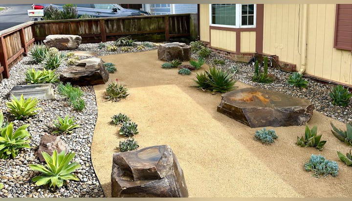 A landscaped garden featuring rocks, succulents, and a sandy path, set in a residential area with a house nearby.