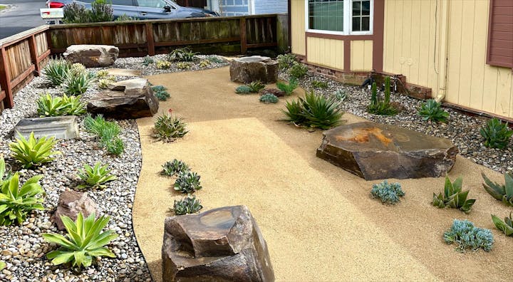 A landscaped garden featuring rocks, succulents, and a sandy path, set in a residential area with a house nearby.