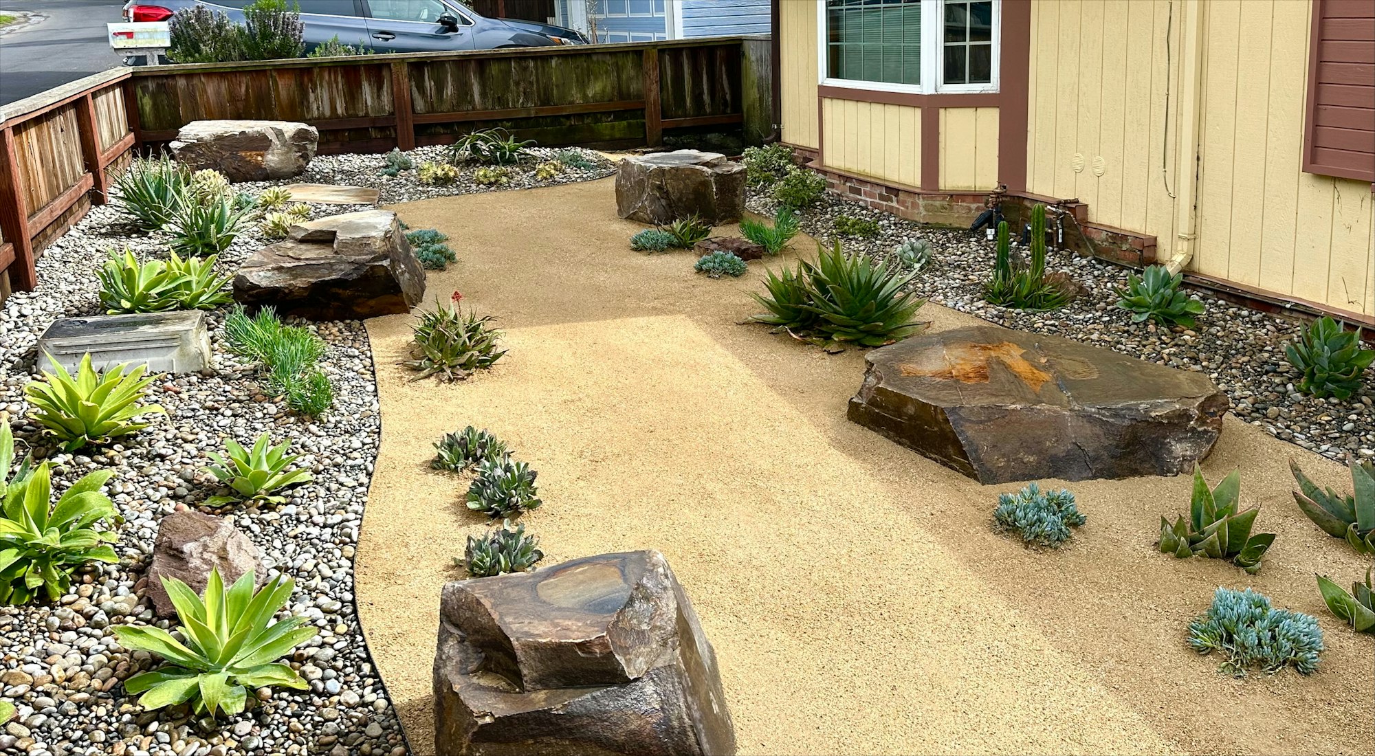 A landscaped garden featuring rocks, succulents, and a sandy path, set in a residential area with a house nearby.
