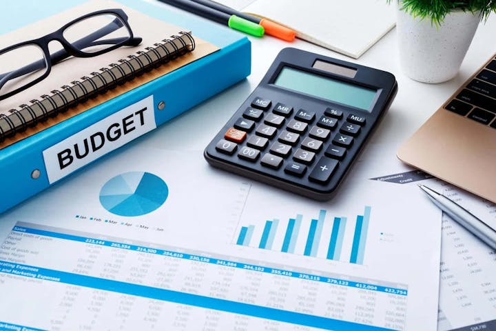 The image shows a budget binder, calculator, glasses, and financial documents with charts and graphs for analysis.