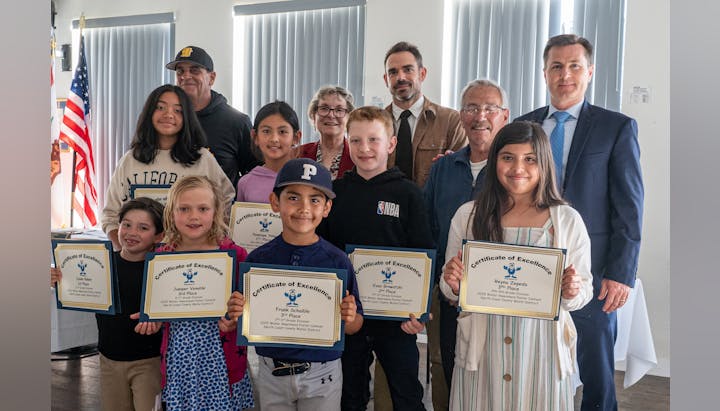 A group of children and adults hold certificates of excellence, posing indoors with flags in the background.