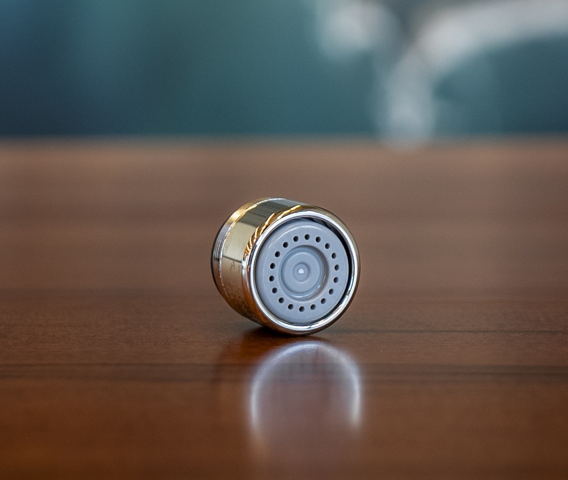 The image shows a small, cylindrical metal object with a round, perforated top, resting on a wooden surface.