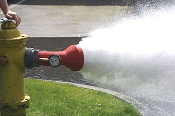 A yellow fire hydrant is spraying water, with a gauge visible on its side, and someone is holding it.
