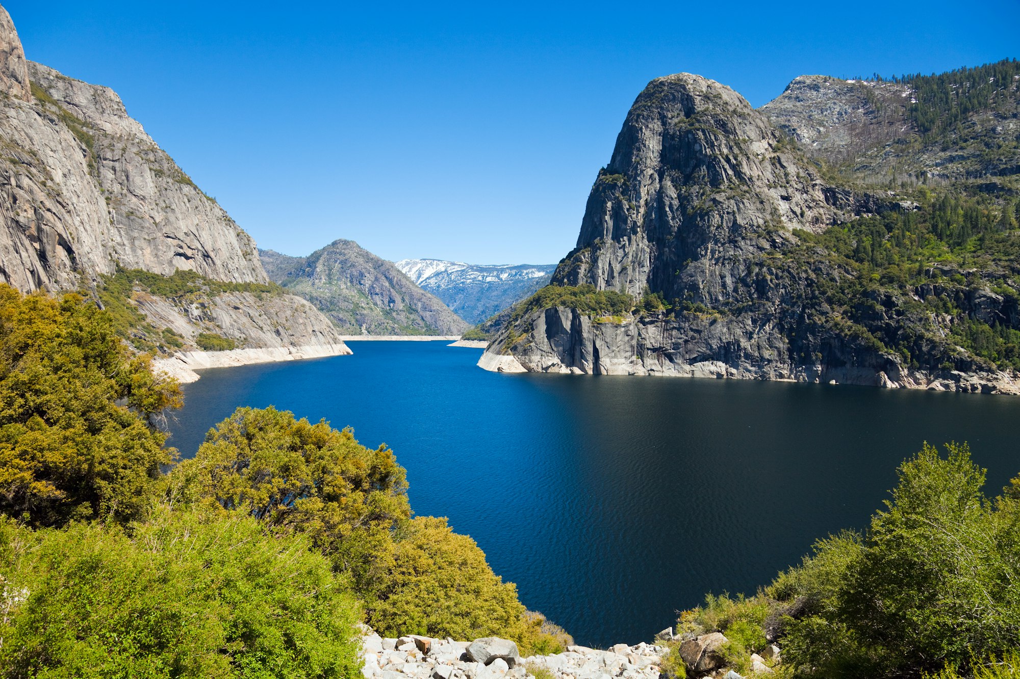 A scenic view of a lake surrounded by mountains and lush greenery under a clear blue sky.
