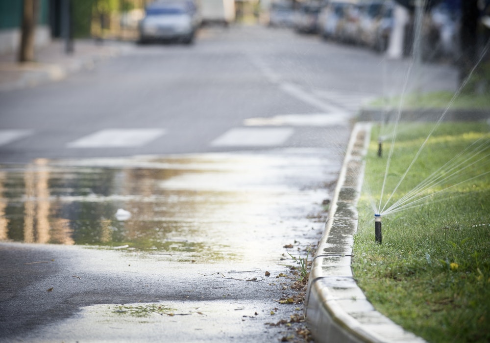A street scene with sprinklers watering grass, reflecting water on the pavement, and cars parked in the background.