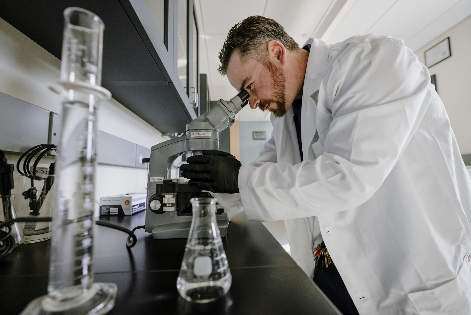 A scientist in a lab coat is using a microscope, surrounded by glassware and lab equipment.
