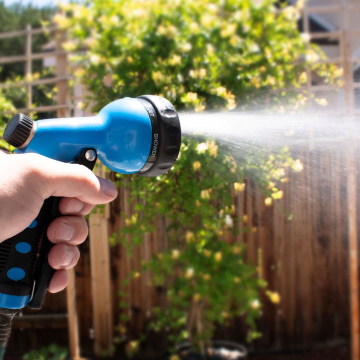 A hand holds a blue garden hose nozzle, spraying water against a backdrop of greenery and a wooden fence.