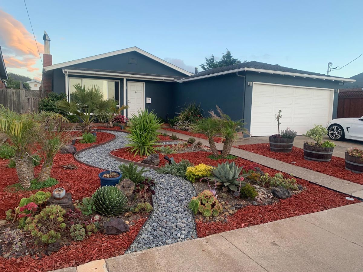 A house with a modern succulent garden, mulched with red bark, a stone path, and a white garage. A white car is parked nearby.