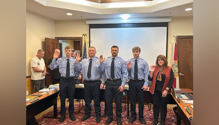 Five people are being sworn in, standing with hands raised. They are dressed in blue uniforms or formal attire in an indoor setting.