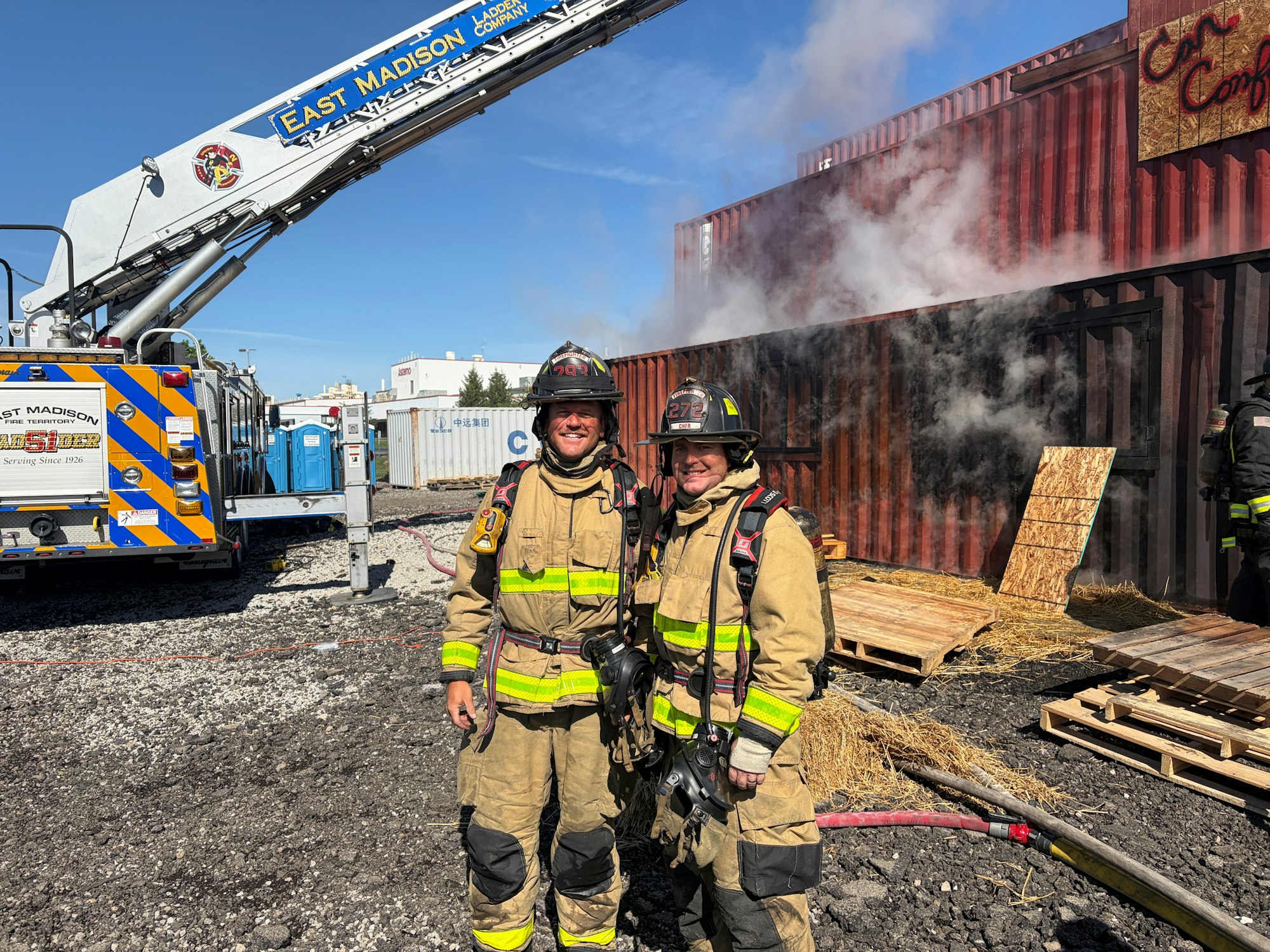 Firefighters pose near a smoke-filled building with a ladder truck in the background, indicating an active fire response scene.