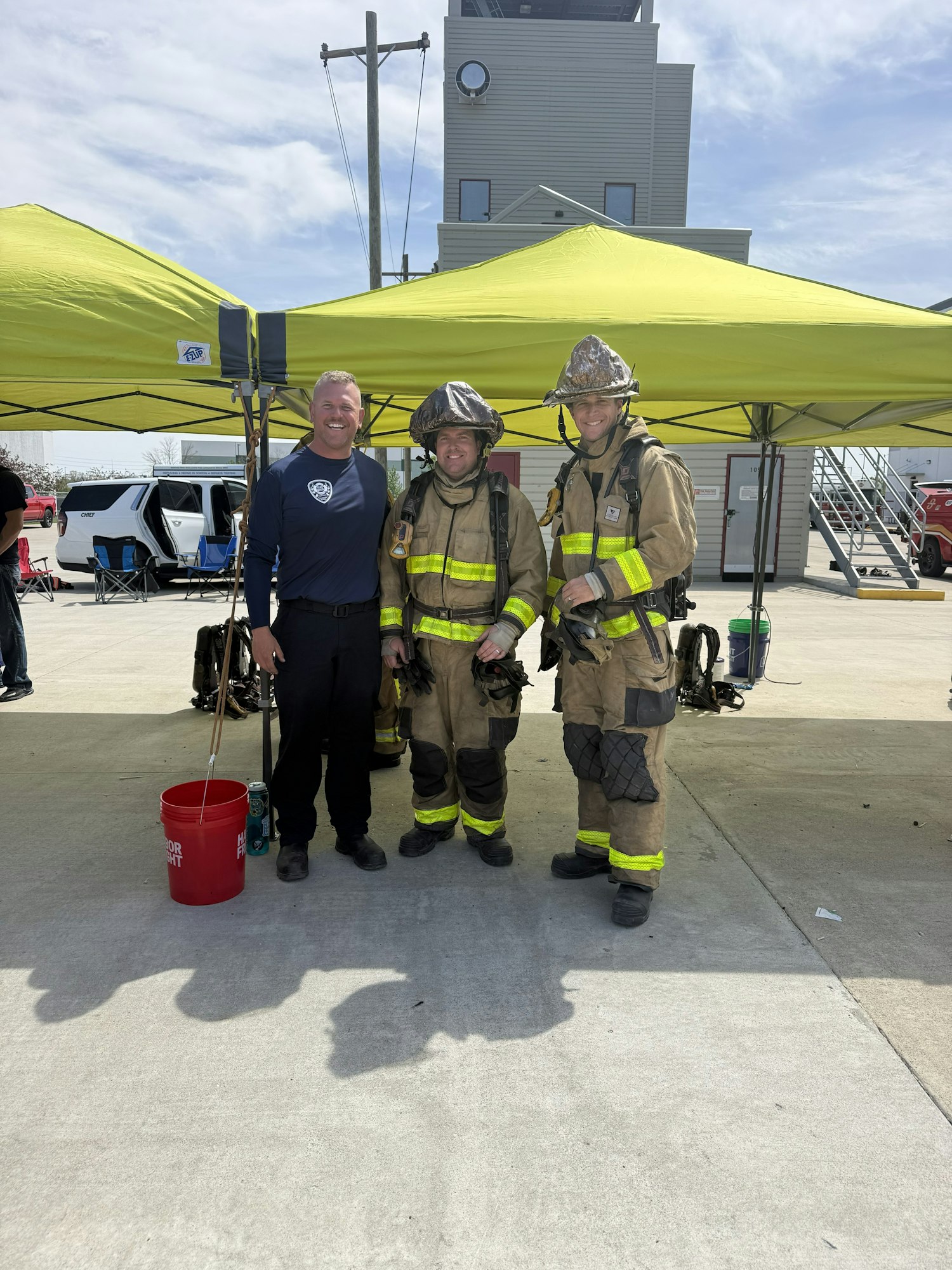 Three firefighters in gear pose with a man in casual clothes under a green tent, with a building and equipment in the background.