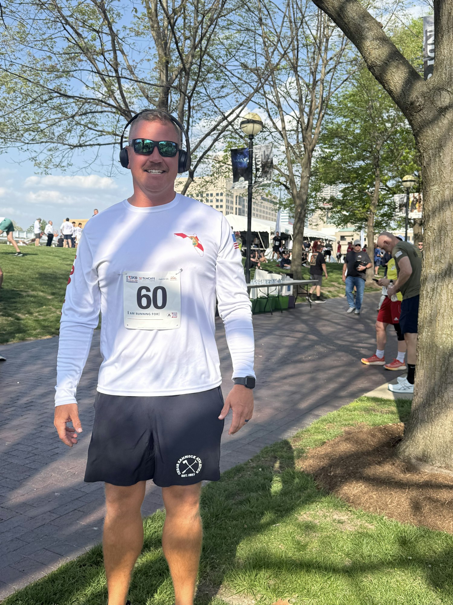 A smiling participant wearing a race number and headphones stands outdoors, surrounded by trees and event-goers.