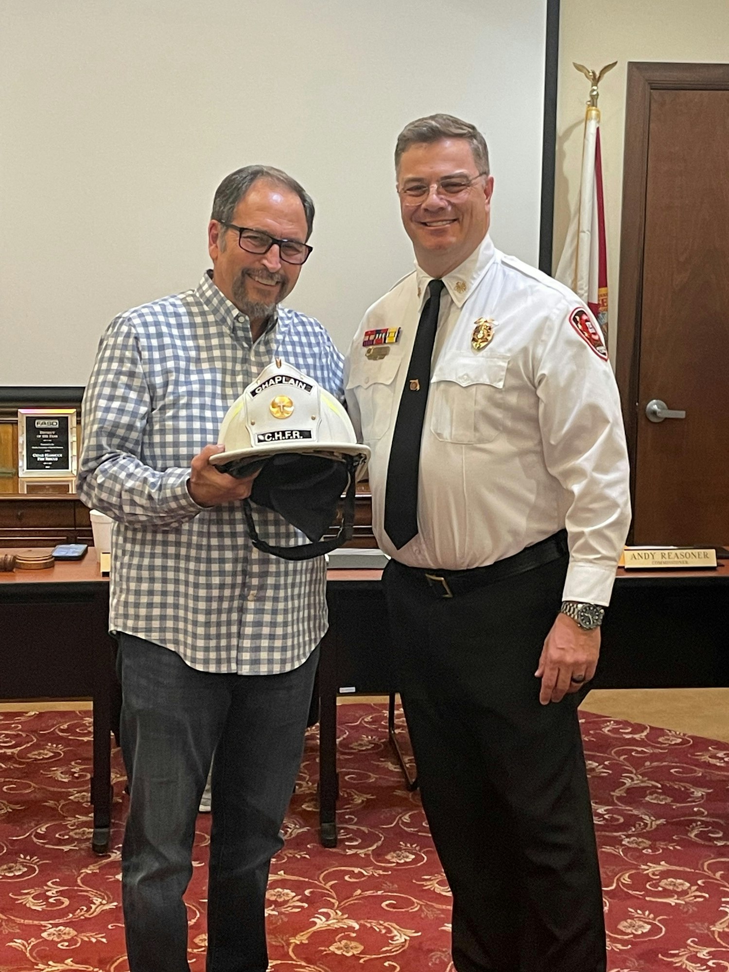 Two men are smiling and posing for a photo; one in a fire chief's uniform is handing a helmet to the other.