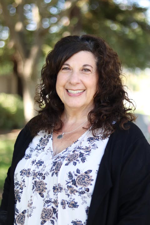 A woman with curly hair smiles, wearing a floral top and a black cardigan, against a blurred outdoor background.