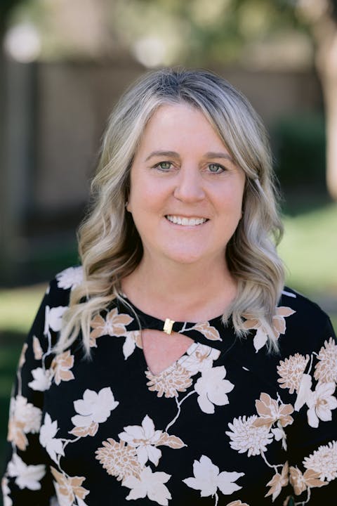 A woman with wavy blonde hair is smiling, wearing a black floral dress, in a green outdoor setting.