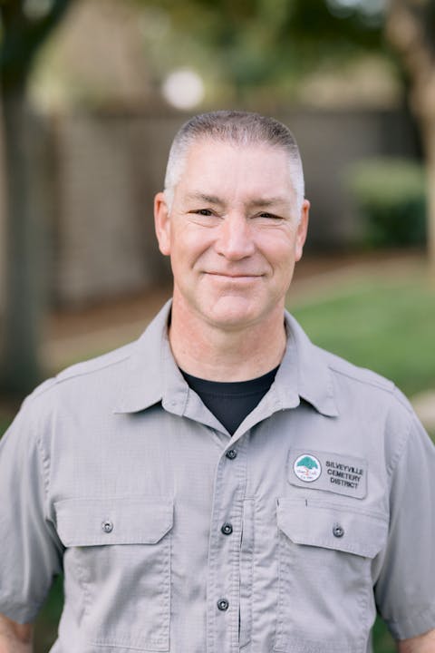 A man with short hair, wearing a gray shirt with a badge from the Silveyville Cemetery District, smiles outdoors.