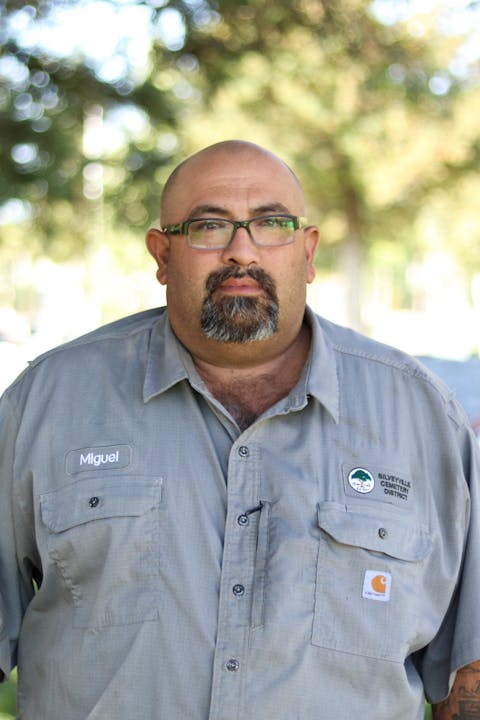 The image shows a man named Miguel wearing a uniform with a cemetery district badge, standing outdoors in a natural setting.