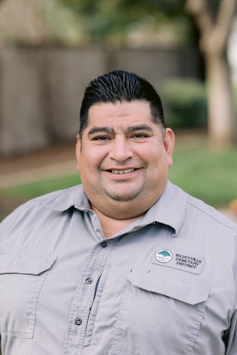 A smiling man wearing a gray shirt with a badge from the Silveyville Cemetery District stands outdoors.
