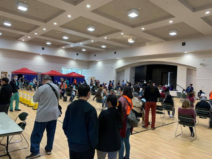 The image shows a large indoor space with many people seated, likely at an event or gathering, with tents and signs present.