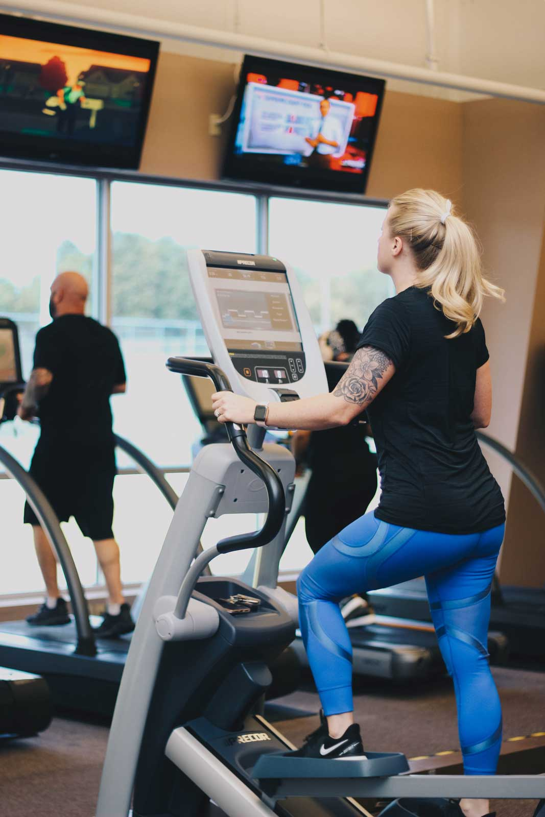 A gym scene with two people exercising on ellipticals, watching TV screens. One person has tattoos and wears blue leggings.