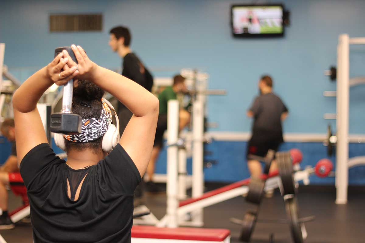 A person with headphones holds a dumbbell above their head in a gym, with others working out in the background.