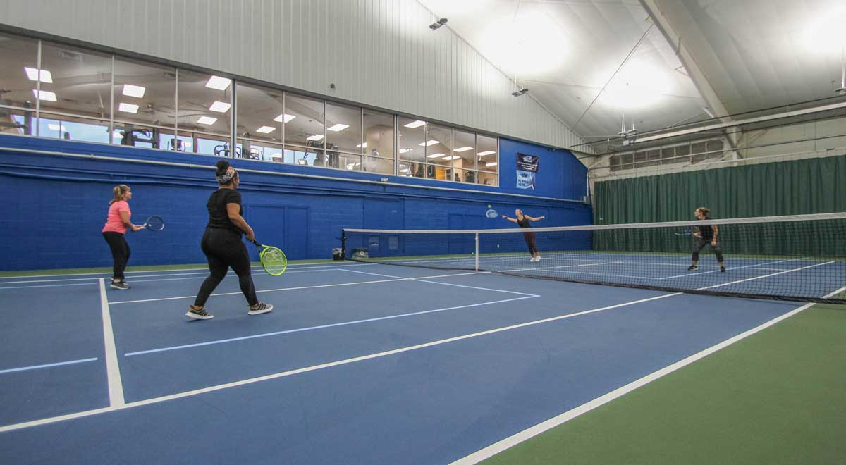 Three people are playing tennis indoors, with a blue court and nets in view. The surrounding area is bright and spacious.