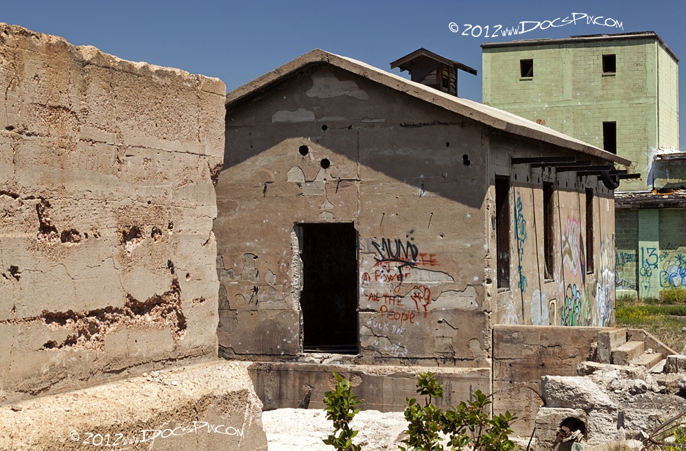 The image shows abandoned structures with visible graffiti, surrounded by natural growth and a clear blue sky.