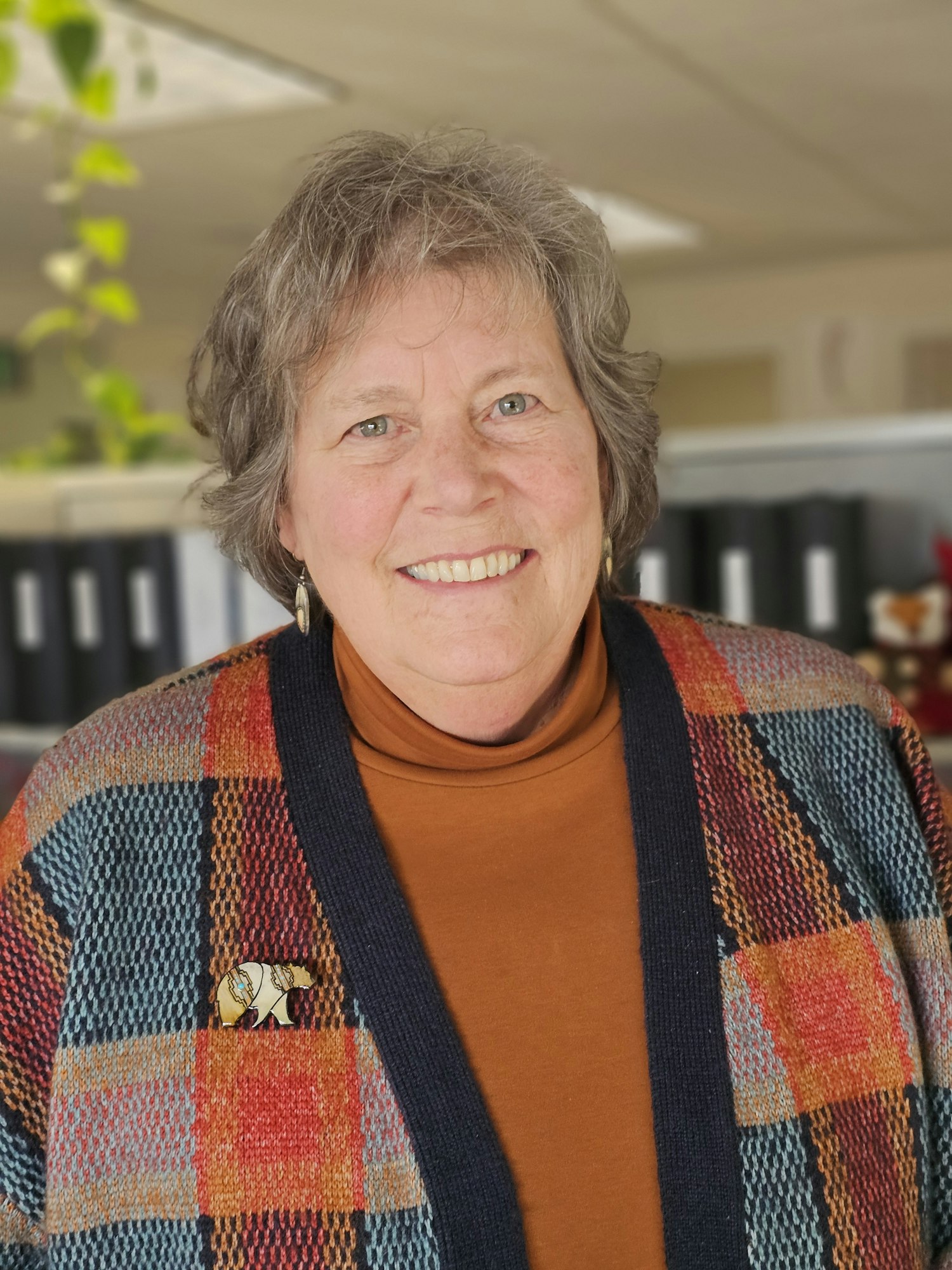 A person smiling, wearing an orange turtleneck and a colorful plaid sweater, standing indoors.