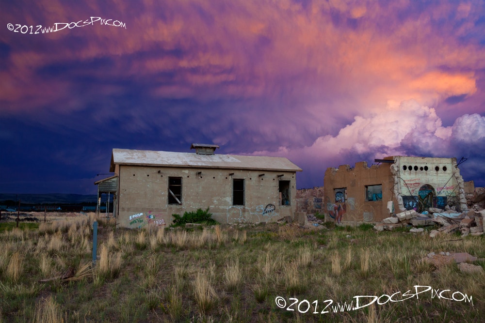 The image features two abandoned buildings amidst tall grasses, set against a dramatic, multicolored sky at sunset.