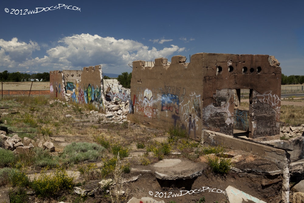 The image shows the remains of a building with graffiti amidst a grassy field under a clear blue sky.