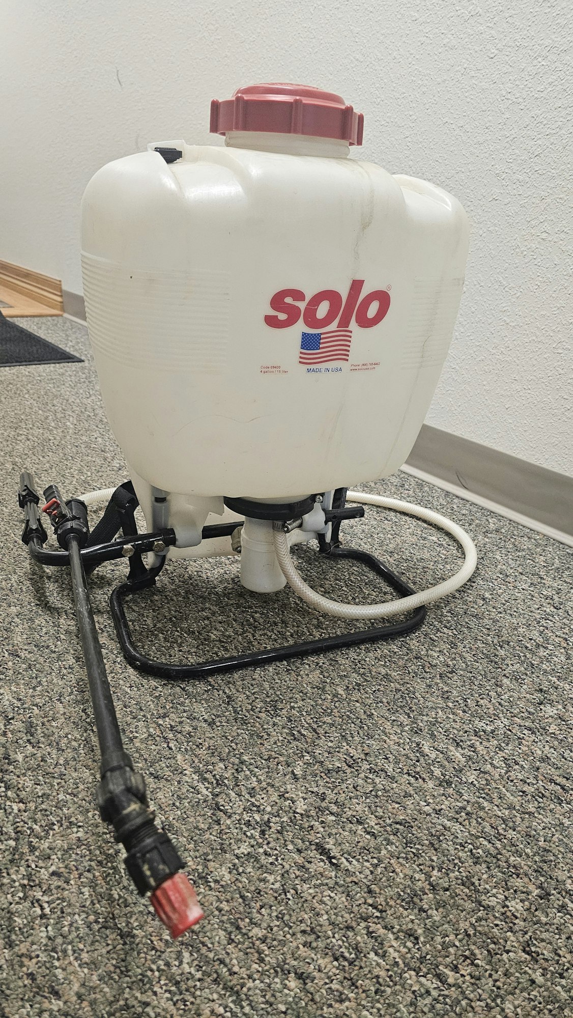 A backpack garden sprayer with a white tank, hose, and spray wand, titled against a wall.