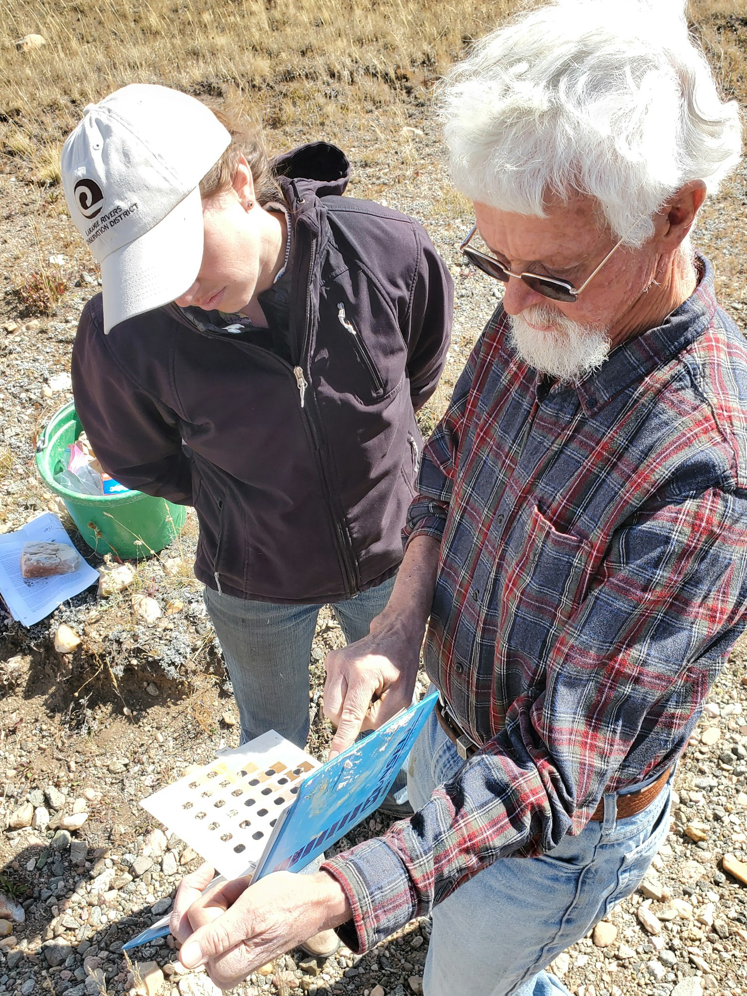 Two people outdoors examining a guidebook, wearing casual clothes and standing on rocky ground.