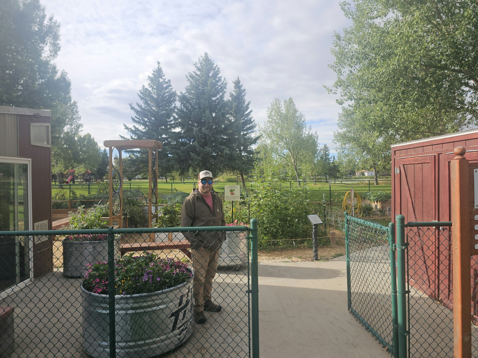 A person stands near a garden with flowers in large pots, surrounded by greenery and trees.