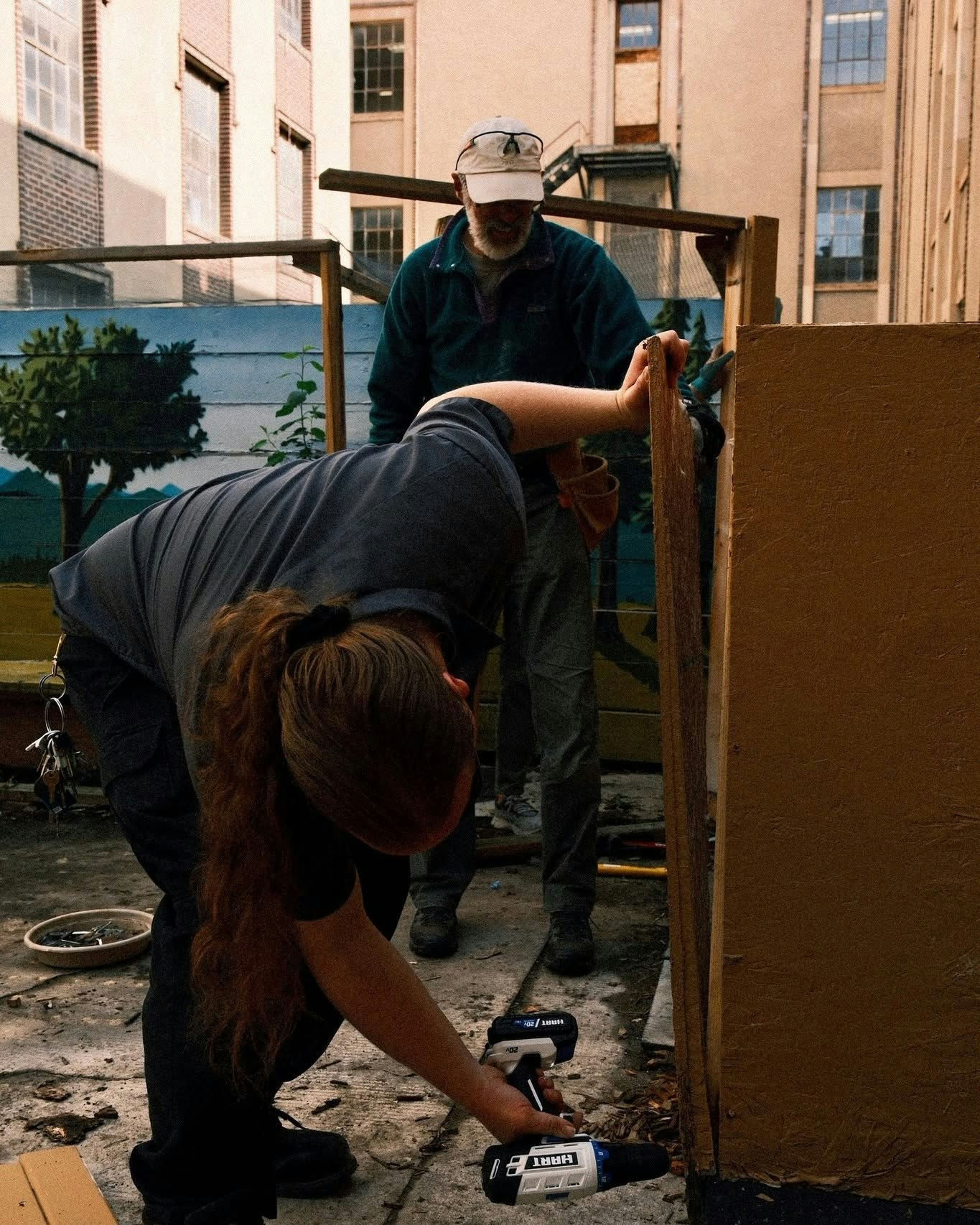 A person is using a power tool to work on a project while another person observes in a courtyard setting.