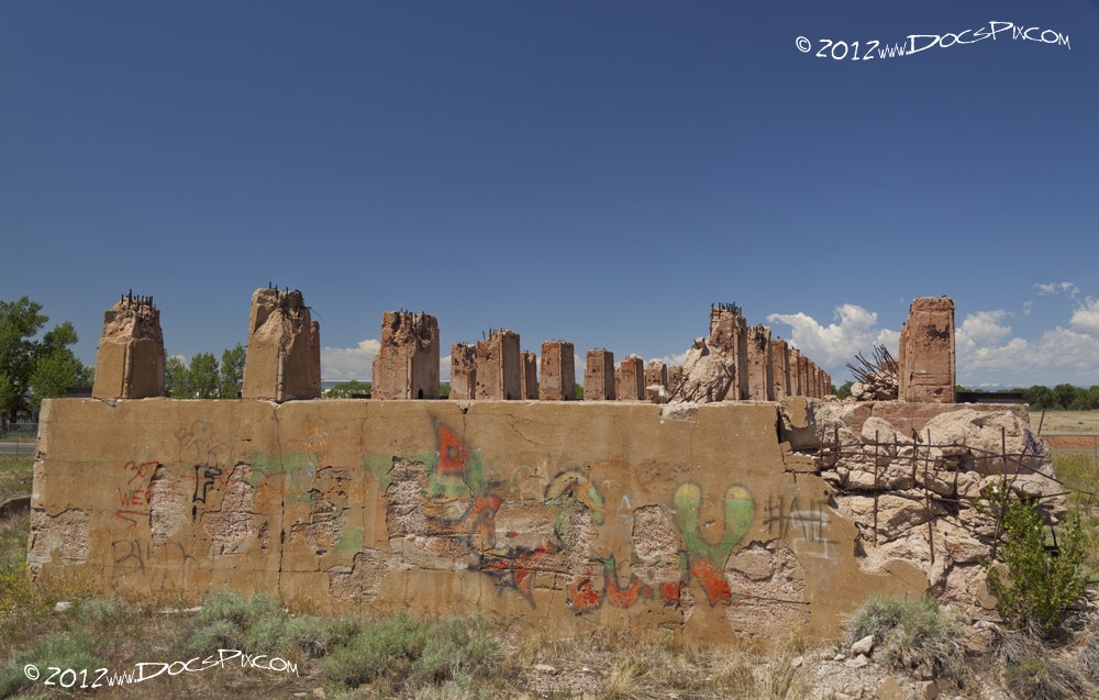 The image shows the remnants of a ruined structure with weathered walls, partially standing columns, and graffiti, set against a blue sky.