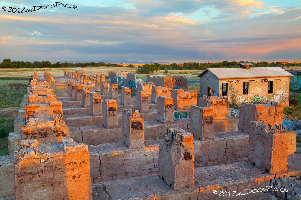 The image shows an expansive area of weathered, graffitied ruins and a small building, with a picturesque sky above.