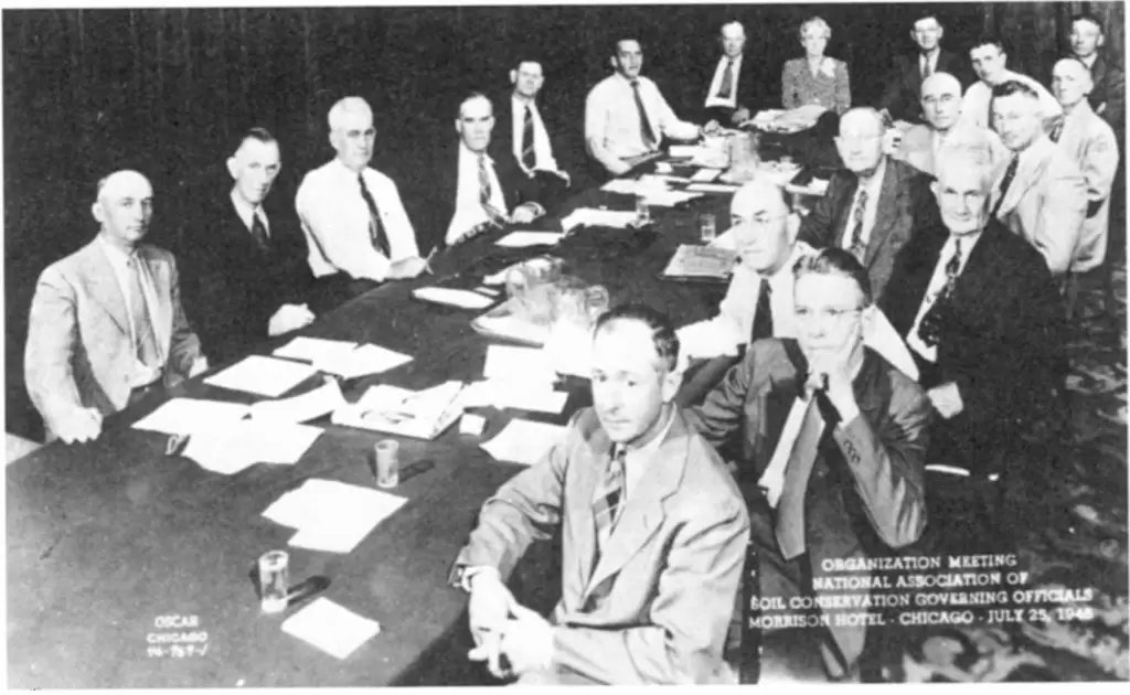 Black and white historical photo of a group of men seated around a large conference table, likely from the mid-20th century. Most are wearing suits and ties, and there are documents and glasses of water on the table.