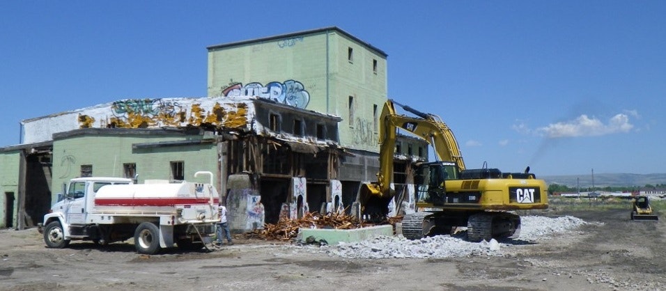 A demolished building with heavy machinery and a truck in an industrial area, surrounded by debris and graffiti.