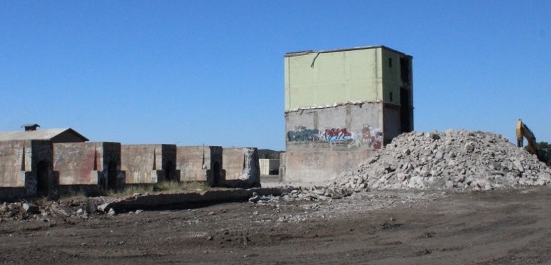 An abandoned, partially demolished building with graffiti, surrounded by rubble and construction equipment under a clear blue sky.