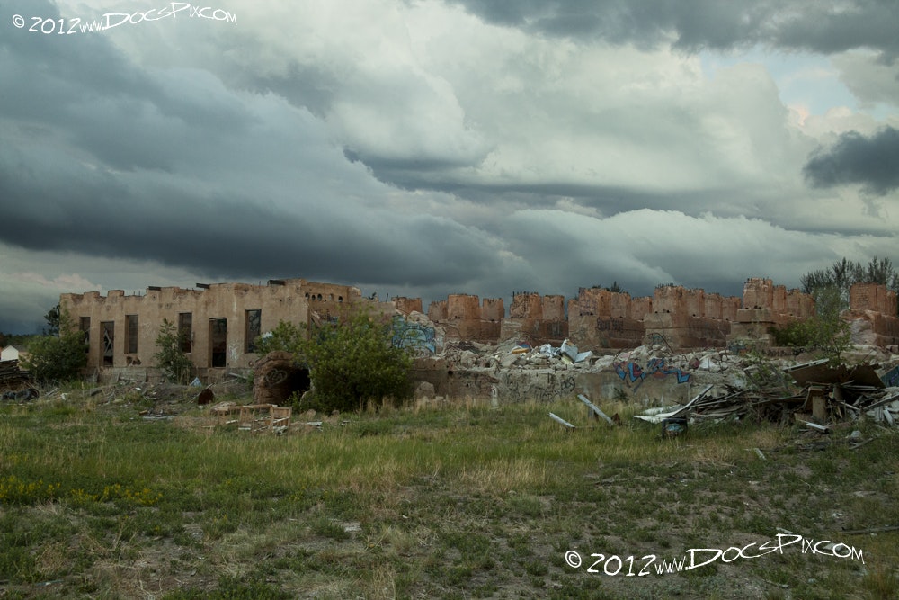 The image shows an abandoned building in ruins, surrounded by overgrown grass and debris, under a cloudy sky.