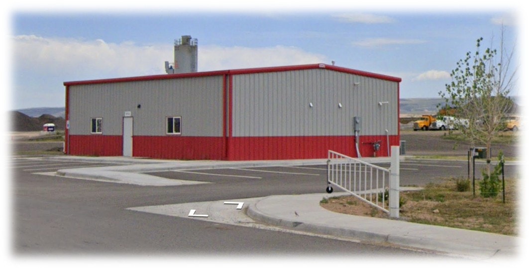 A red and gray industrial building with a fenced parking area and a visible silo in the background.