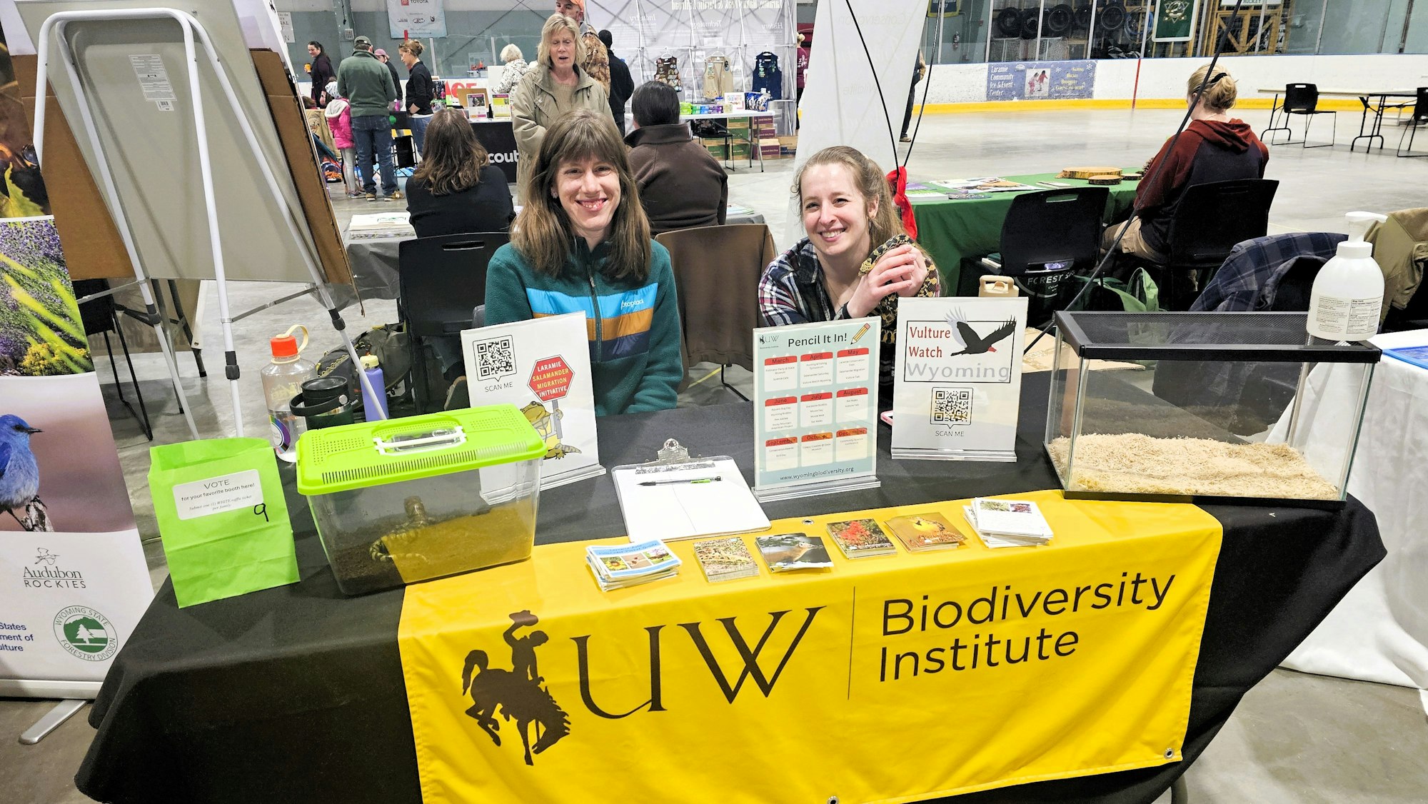 Two people at a Biodiversity Institute booth with educational material, a turtle in a container, and a terrarium.