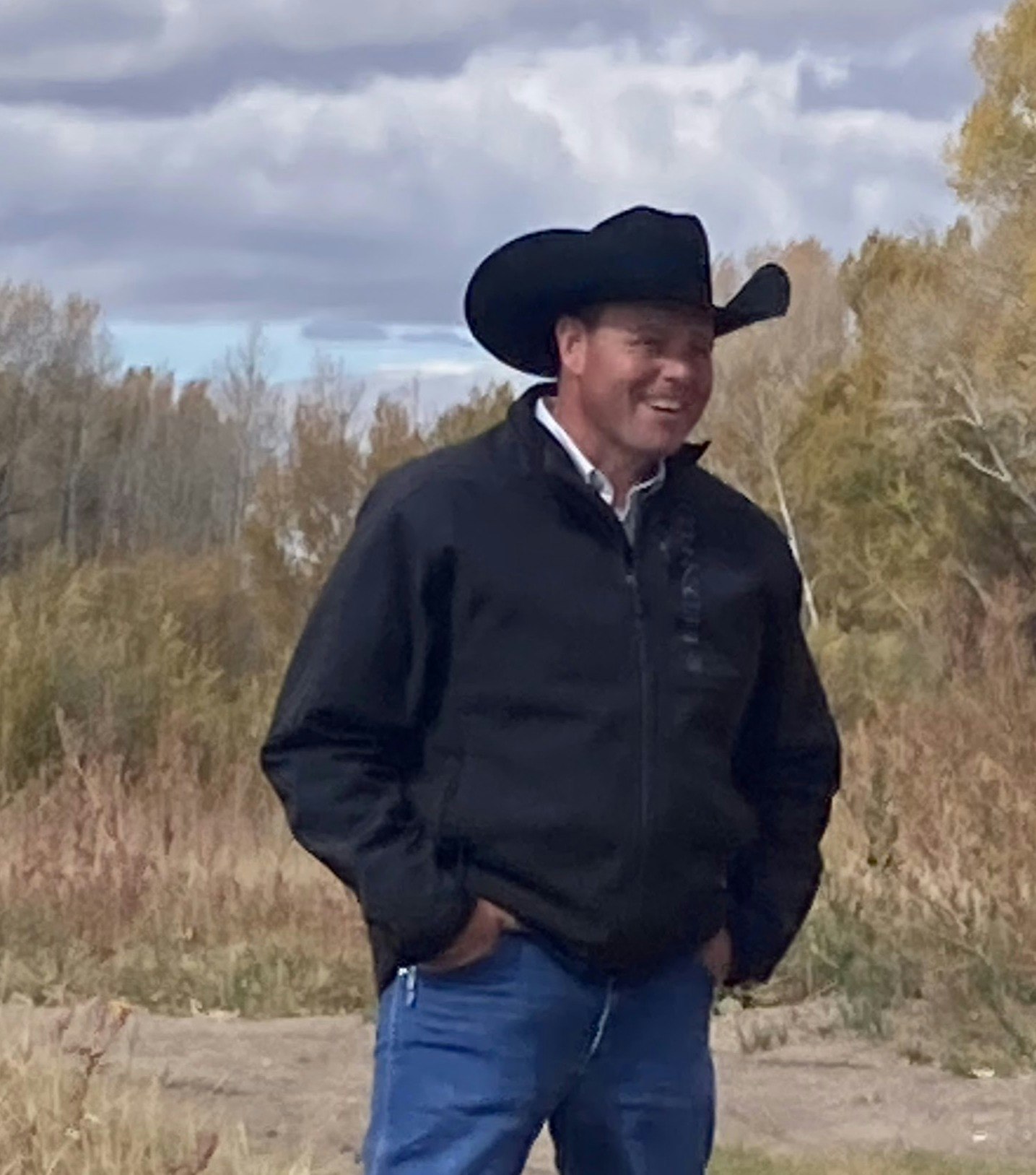 A person wearing a black cowboy hat and jacket, smiling, standing outdoors with a cloudy sky and trees in the background.
